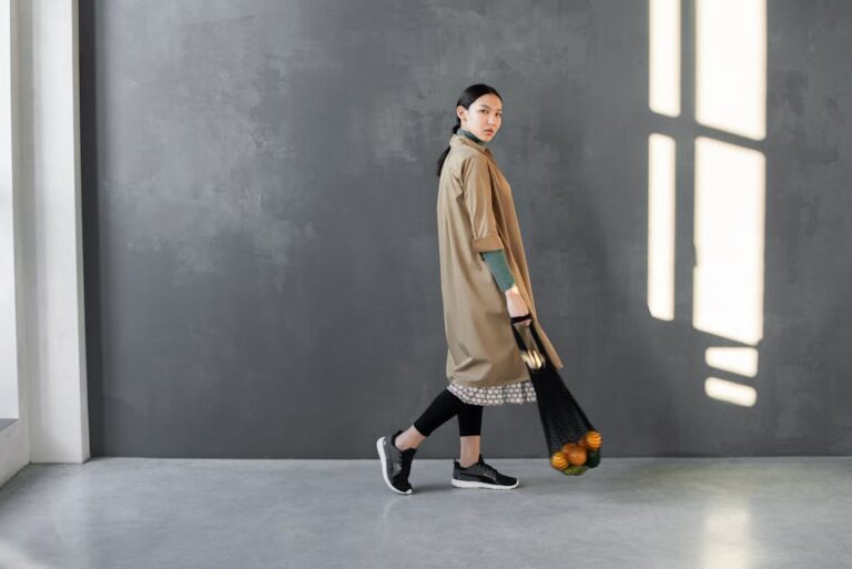 Stylish woman posing indoors with a net bag of fruits, wearing a trendy oversized coat.