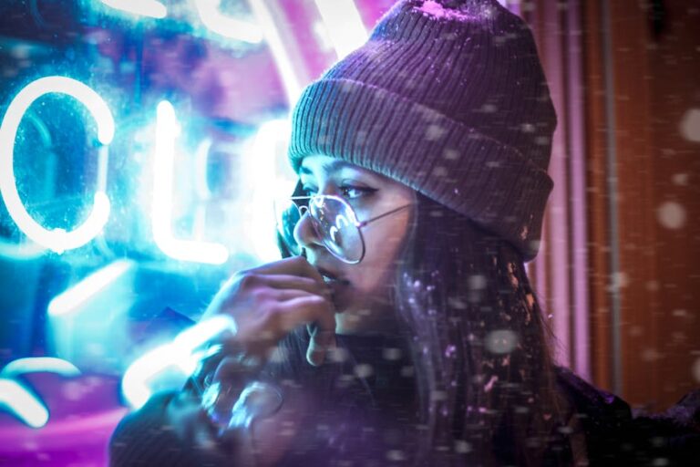 A woman in a beanie and glasses stands by neon lights during a snowy night.