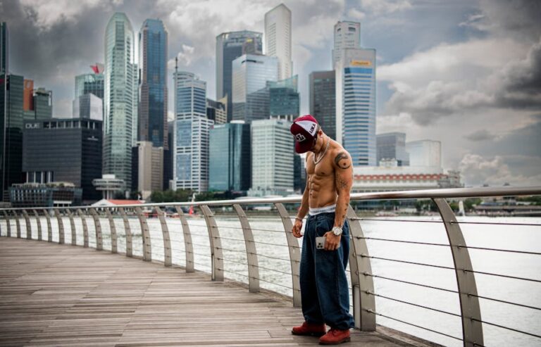 Man in casual attire stands on boardwalk with city skyline in background.