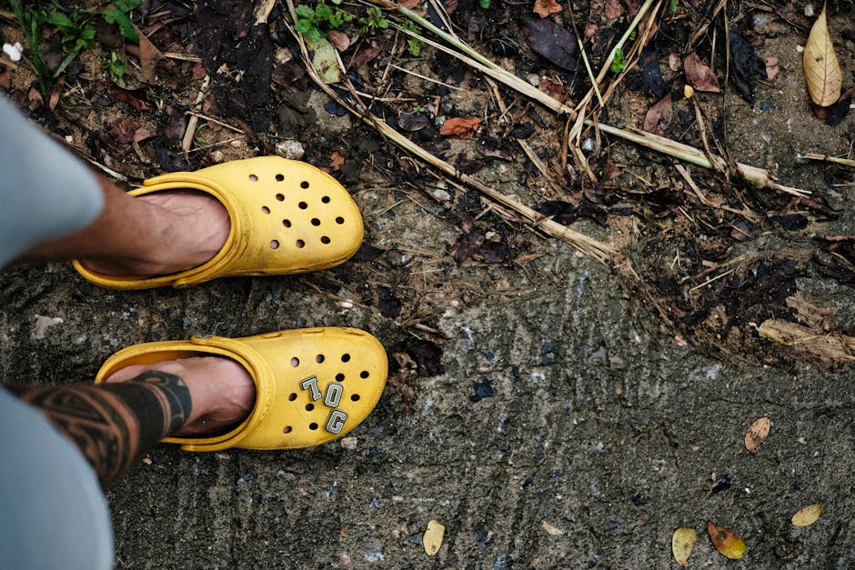 Man in yellow Crocs with tattoo on foot standing on earthy path with dried leaves.