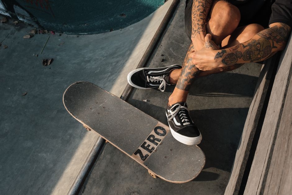 Tattooed skateboarder in black shoes sitting at a skatepark with skateboard. Capturing street style and lifestyle vibe.