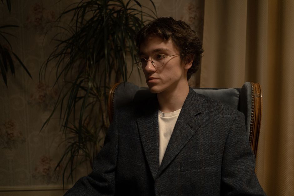 A man in a blazer and eyeglasses sitting seriously in an armchair, surrounded by house plants and curtains.