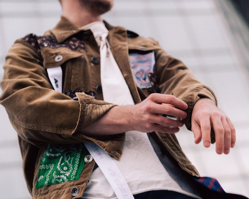 Stylish man in a brown jacket with patches, adjusting sleeves in an urban setting.