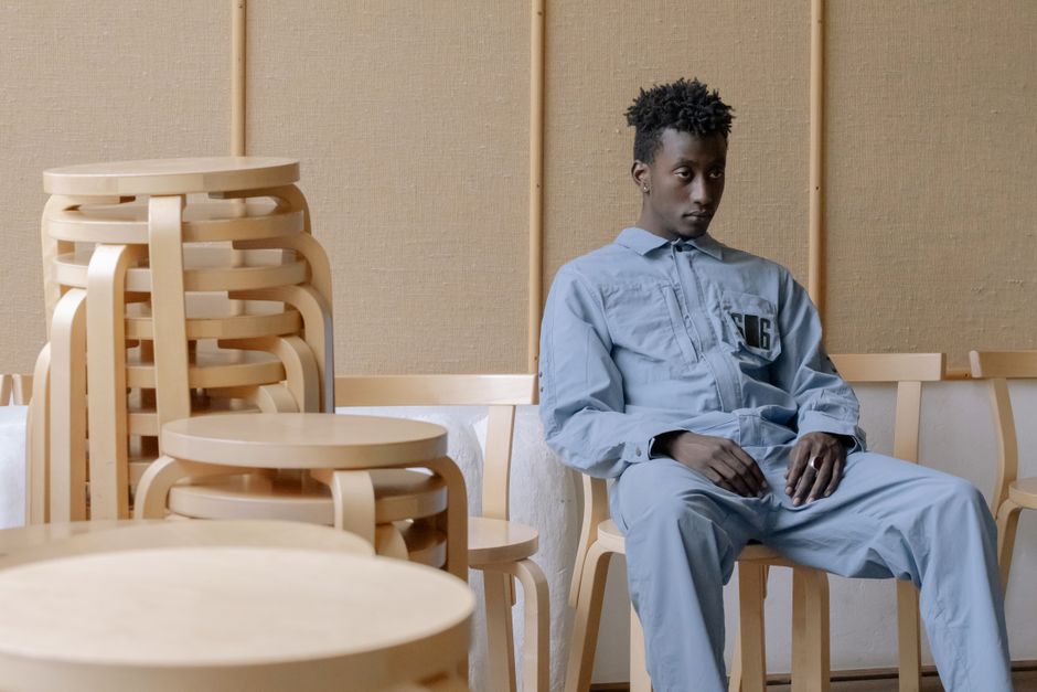 A young man in a blue jumpsuit sitting in a room with stacked wooden chairs.